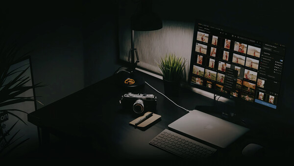 Photographer's desk setup with camera, laptop displaying image library, and monitor in moody side lighting.