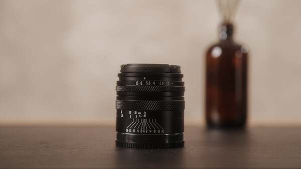 Black vintage lens with depth of field markings sits on wooden surface with brown glass bottle blurred in background.