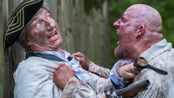 Two men in period costume engaged in a physical altercation near a wooden structure.