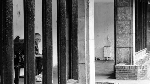 Person seated behind vertical wooden columns creating layered geometric composition in black and white.