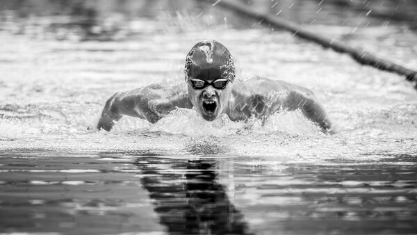 Competitive swimmer performing butterfly stroke in rain, captured mid-stroke with water splashing around their face and goggles.
