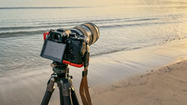 Professional camera on tripod mounted at beach during golden hour, facing ocean waves and shoreline.