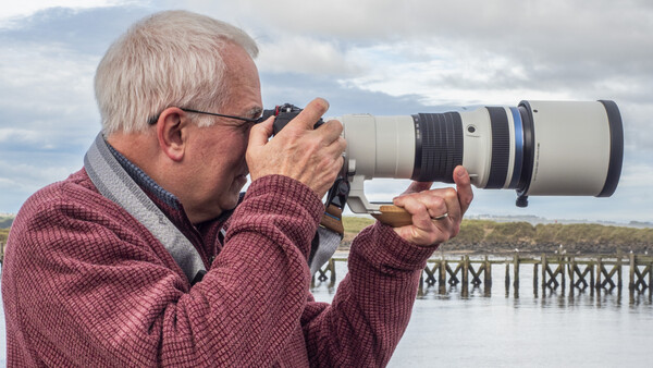 Older photographer holding a telephoto lens up to his eye while standing at a railing overlooking water.