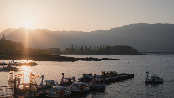Golden sunrise light illuminates a calm alpine lake with moored boats in the foreground and mountains silhouetted across the water.