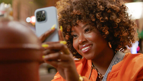 Woman in orange jacket holding white smartphone, smiling at camera in warm indoor lighting.