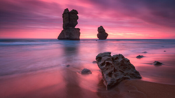 Rocky sea stacks silhouetted against a vibrant pink and purple sunset reflected in calm shallow water.