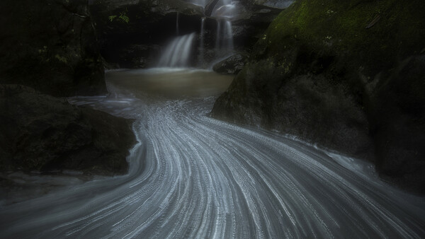 Long-exposure photograph of a flowing stream with silky water patterns curving around moss-covered rocks and a waterfall in the background.