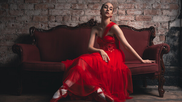 Woman in a red tulle gown seated on a burgundy velvet sofa against an exposed brick wall.