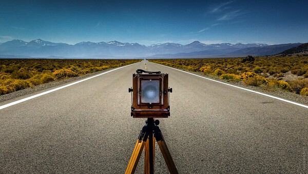 Large format camera on tripod centered on a straight desert highway with mountains in the distance.