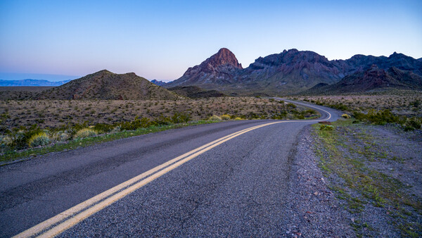 Desert highway with double yellow lines curving toward distant mountains at dusk.