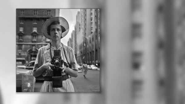 Woman in wide-brimmed hat holding a large format camera on an urban street.
