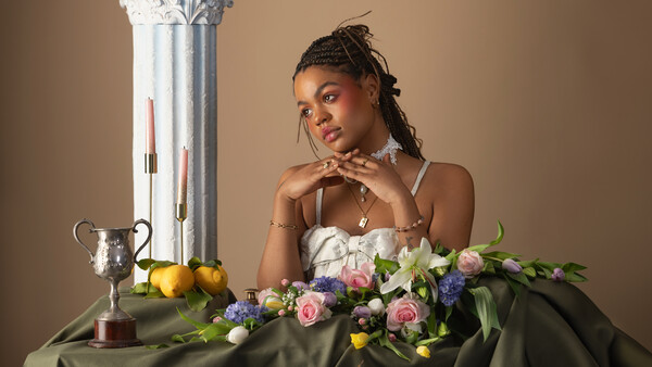 Woman posing with floral arrangement and decorative still life elements against neutral backdrop.
