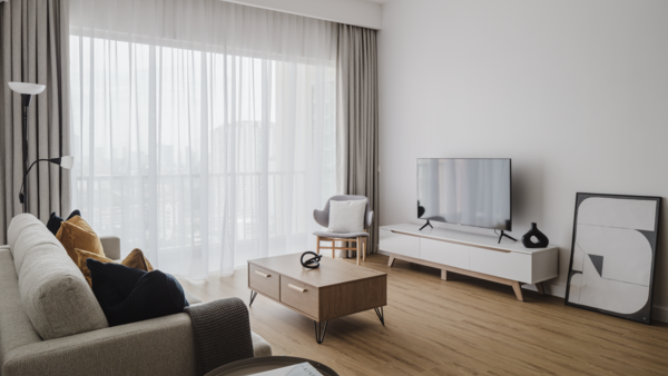 Modern living room with light wood flooring, neutral upholstered furniture, and large sheer curtains filtering natural light from floor-to-ceiling windows.