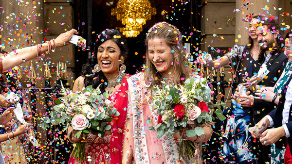 Two people in traditional wedding attire smiling amid falling colorful confetti.