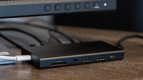 Docking station with multiple ports connected to a laptop, sitting on a wooden desk next to a monitor.