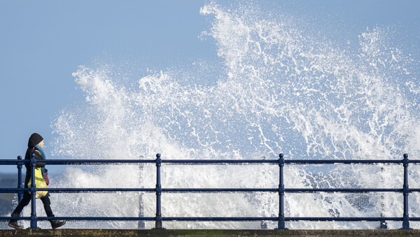 Person in yellow safety vest standing at railing as large wave crashes against barrier.