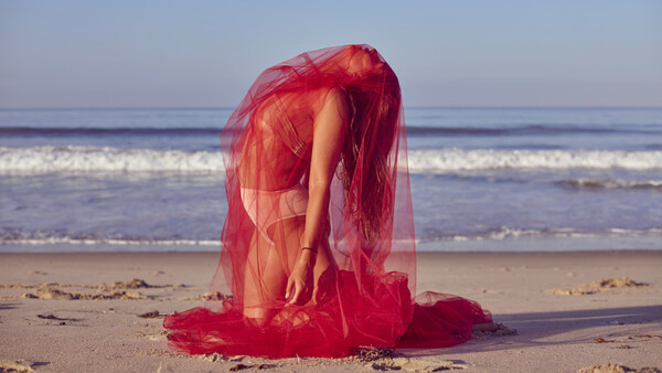 Person wrapped in sheer red fabric on a beach, bent forward with arms extended toward the sand.