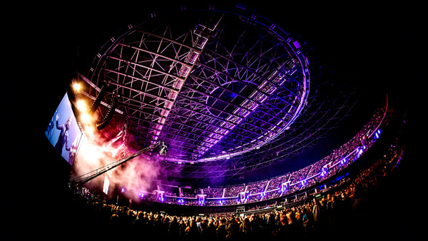 Large concert venue interior with purple stage lighting and crowd silhouettes against bright stage effects.