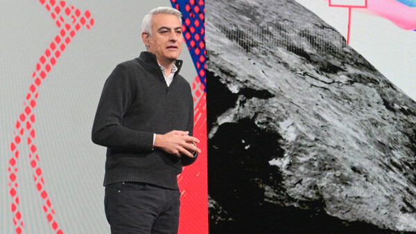 Speaker gesturing while presenting on stage with abstract geometric graphics and a black and white mountain photograph displayed on large screens behind him.