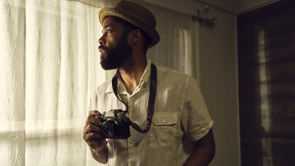 Photographer holding vintage film camera while looking out window with natural sidelight.
