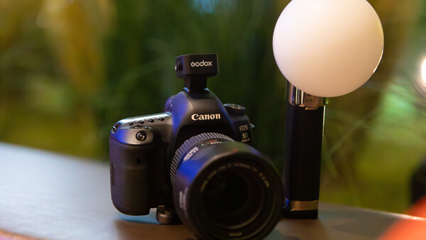Canon DSLR camera with Godox flash and diffuser dome mounted on top, set against blurred outdoor background.