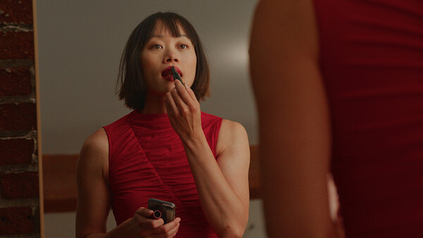 Woman in red sleeveless turtleneck applying lipstick while looking in mirror.