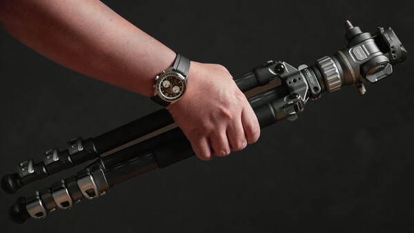 Photographer's hand gripping a telephoto lens with a watch visible on the wrist against a dark background.
