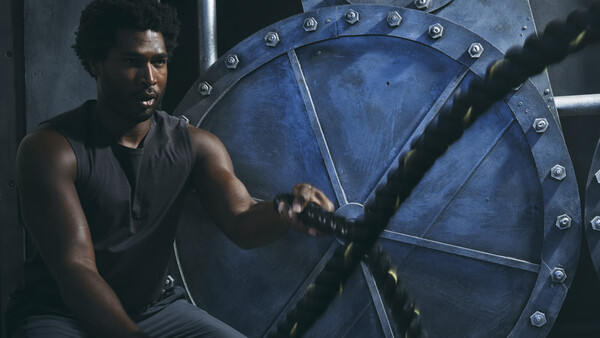 Man in dark tank top sits against large blue industrial gear wheel with metal studs and rivets.