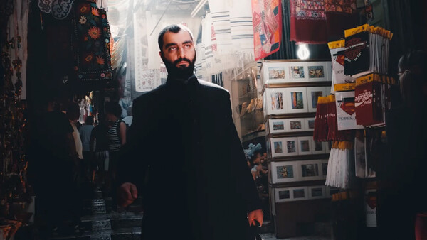 Bearded man in dark coat walking through narrow marketplace corridor lined with merchandise displays.