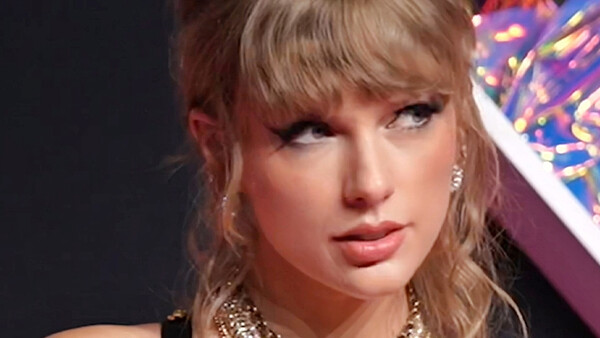 Close-up portrait of a woman with blonde hair and bangs, wearing dramatic makeup and gold chain necklace.