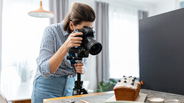 Photographer holding a professional camera with telephoto lens on a tripod while composing a food still life shot.