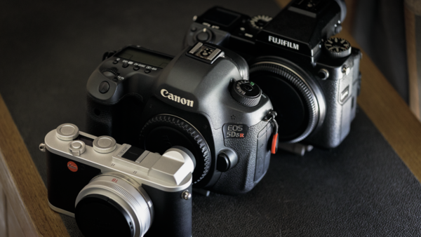 Three digital cameras arranged on a dark surface: a Canon EOS 5D Mark IV, a Fujifilm X-T4, and a compact mirrorless camera in silver.