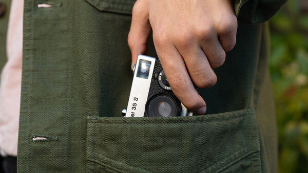 Hand holding a compact rangefinder camera with white body and black lens protruding from olive cargo pants pocket.