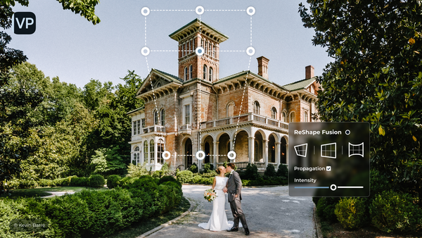 Couple in wedding attire posing in front of a brick Victorian mansion with a tower, surrounded by landscaping.