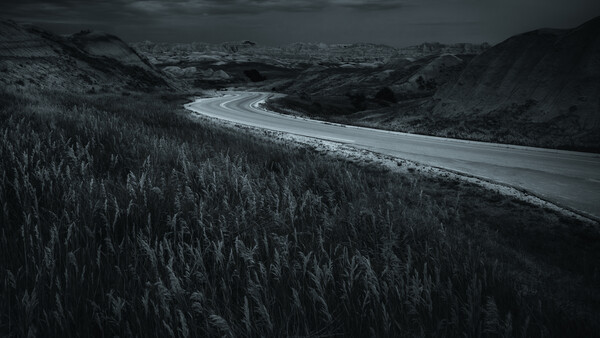 Winding road cutting through dense coniferous forest with mountains in the distance under dark, overcast sky.