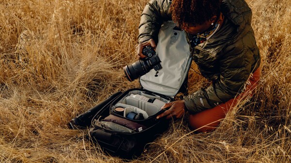 Photographer crouching in dry grass, adjusting a camera backpack with a large telephoto lens visible.