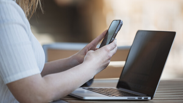 Person holding a smartphone while working at a laptop outdoors in natural daylight.