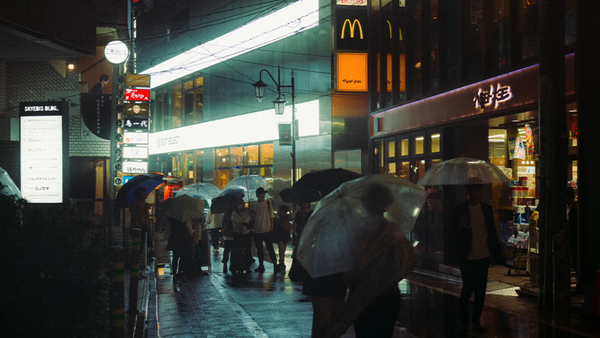 Rainy urban street at night with pedestrians holding umbrellas beneath illuminated storefronts and signs.