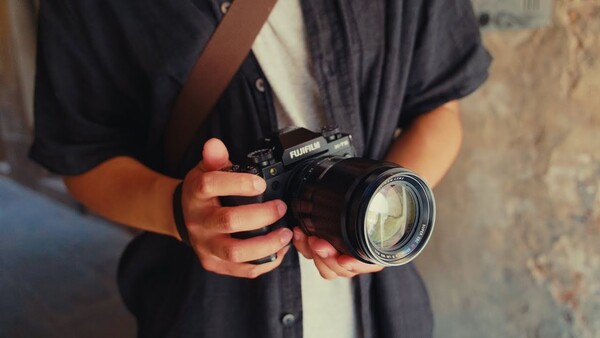 Photographer holding a black Fujifilm camera with a telephoto lens against their chest.