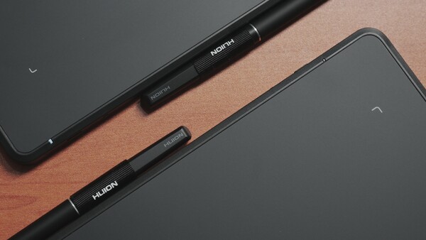 Two black stylus pens labeled with text, arranged diagonally on a wooden surface next to tablet devices.