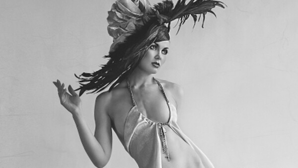 Woman in halter top with dramatic feathered headpiece, shot against neutral background in black and white.