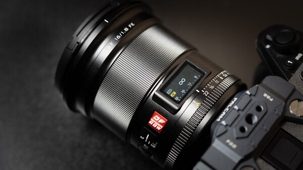 Close-up of a mirrorless camera lens with manual focus ring and autofocus controls mounted on a camera body against a dark surface.