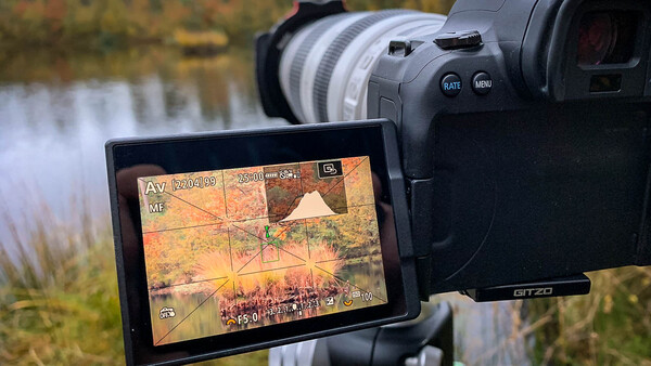 External monitor displaying video footage with grid overlay and exposure information mounted on professional video camera beside water landscape.