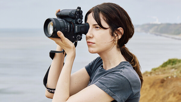 Female photographer holding a DSLR camera with telephoto lens, positioned on a coastal cliff overlooking the ocean.