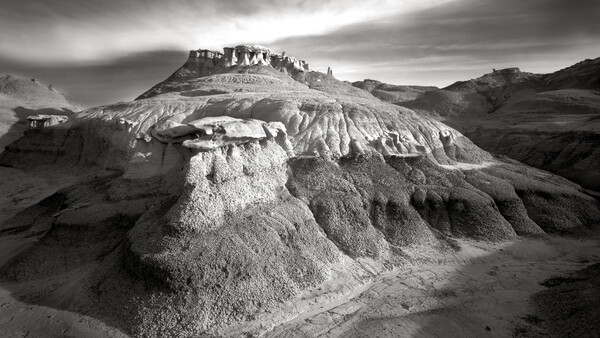 Dramatic black and white landscape of layered, eroded badlands with structures atop the highest ridge.