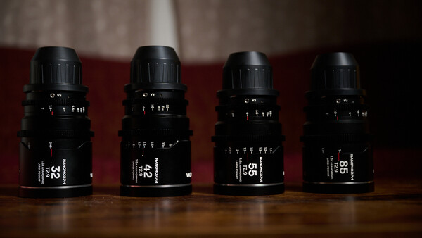 Four black cinema lenses arranged in a row on a wooden surface against a burgundy backdrop.