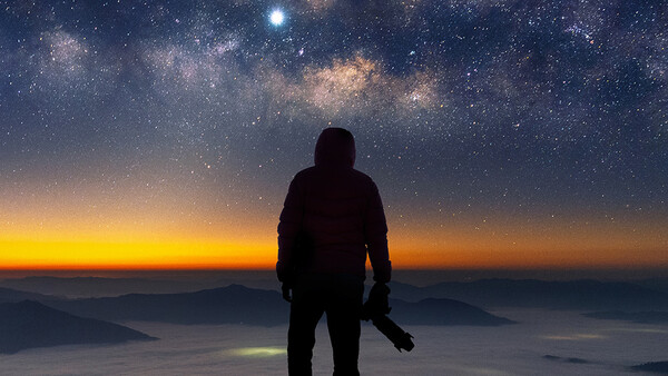 Silhouetted photographer standing on a mountain overlook at sunset, facing a star-filled sky with the Milky Way visible above the horizon.