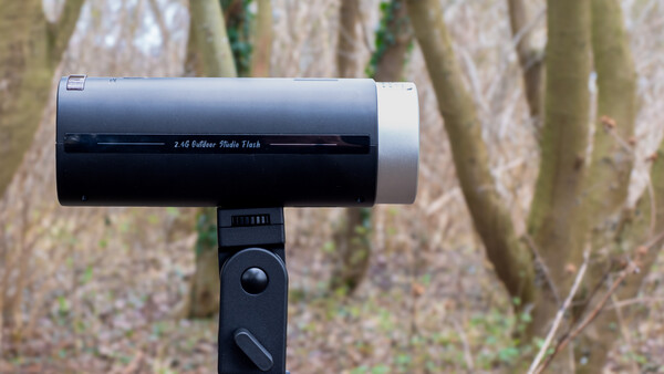 A telephoto lens mounted on a tripod head, positioned against a blurred forest background with tree trunks and vegetation.