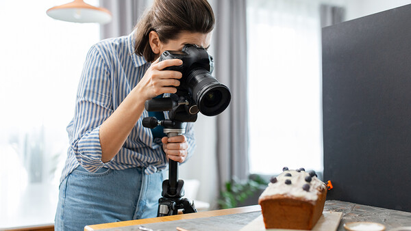 Female photographer holding a mirrorless camera with telephoto lens mounted on a tripod, shooting a styled food setup.