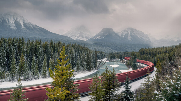Snow-covered mountain landscape with coniferous forest and red train curving through valley.
